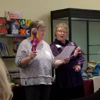 A woman holding a puppet in one hand reads a poem in her other hand, while a second woman looks at the poem
